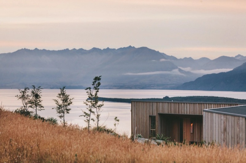 A timber lodge on a golden hillside above a lake at dawn, mountains beyond.
